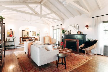 Living room with white vaulted ceilings, wood floors, black fireplace with brick accent, retro space chairs, and red-neutral rug