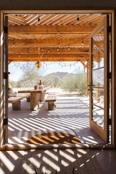 Open French doors, leading to a wood pergola with string lights. A wood picnic table with plants and an ochre pendant light is above.