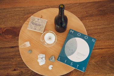 Round wood coffee table with a book titled, 'Moon Lists', a bottle, box of matches, and crystals.
