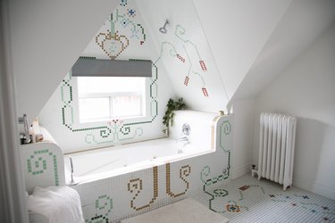 Bathroom with mosaic floral and spiral tile floor, wall, and bathtub. White radiator, pothos, and angular ceiling.