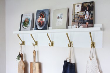 White kitchen wall hangers with gold hooks holding cutting boards, blue apron, and bag of tomatoes on white wall with books resting on top