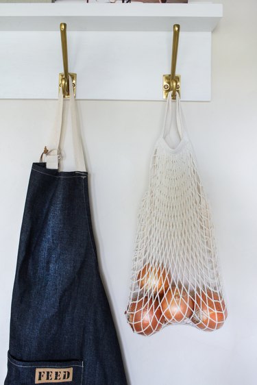 White wall hanger with gold hooks holding blue apron and bag full of tomatoes against white wall
