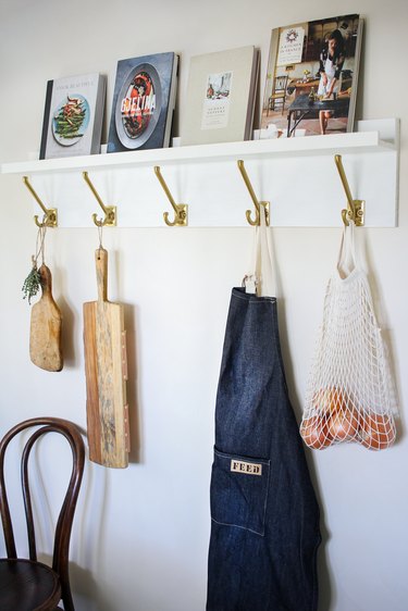 White kitchen wall hangers with gold hooks holding cutting boards, blue apron, and bag of tomatoes on white wall with books resting on top