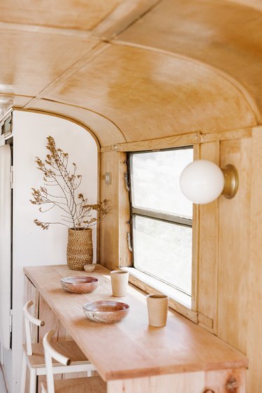 Interior of a cabin, with wood walls, globe sconce, wood table, white stools, basket vase of dried florals, and beige dish ware.