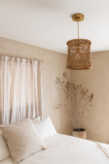 Minimalist bedroom with beige walls, white bedding, beige curtains, boho pendant light, and  dried florals.