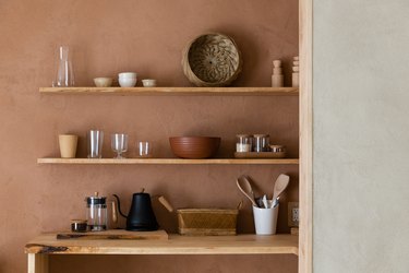 Wood shelves with a round woven bowl, clear glasses, a brown bowl, and containers. Wood table with a French Press and black teapot.