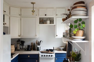Eclectic kitchen with white overhead cabinets, blue lower cabinets, apartment size stove, open shelves, dishes, plants.