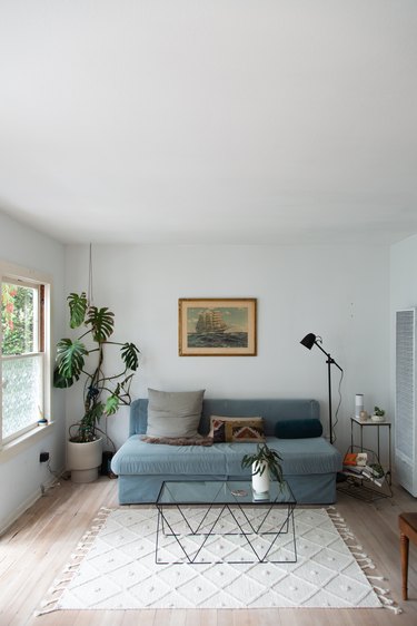 Living room with a gray sofa, glass-metal triangle coffee table, nautical painting, and Monstera plant.