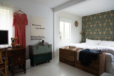Bedroom with Art Nouveau wallpaper, vintage trunks, wood dresser, and wood desk. A poster says, 'All Is Pretty. Andy Warhol.'