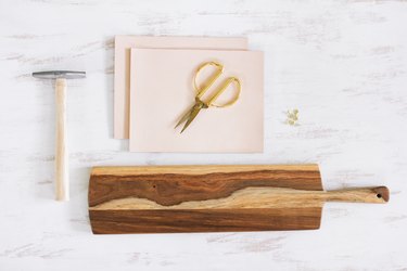a paddle-shaped wooden cutting board, scissors, tacks, a tack hammer, and leather squares