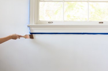 Hand painting white wall along edge of windowsill edged with blue painters tape