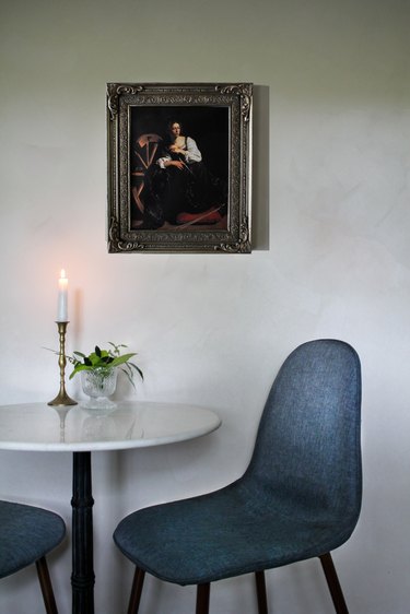Table with lit candle and two blue chairs against white lime-washed wall with framed painting