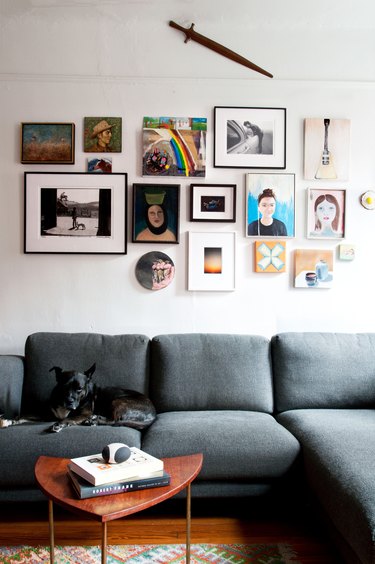 Living room with grey-green couch, dog, coffee table, and white wall with gallery wall and hung sword