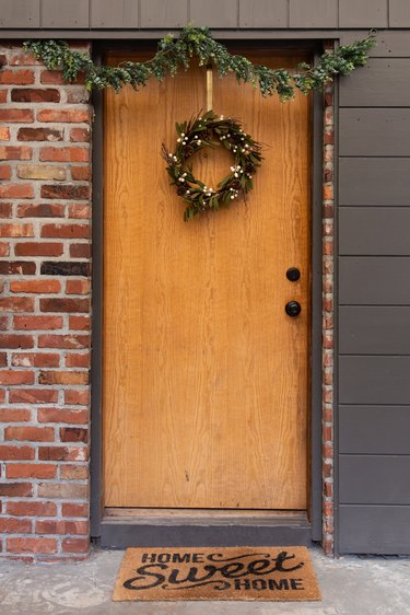 Christmas wreath, holiday wreath on front door with garland, and doormat that says, 'Home Sweet Home'.