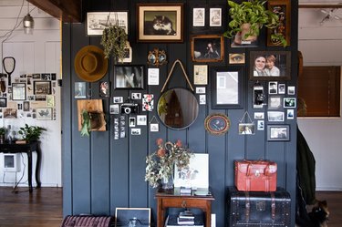 A black wall with wainscoting. Decorated with plants, eclectic art, a mirror, and a hat. Below, vintage trunks and furniture.