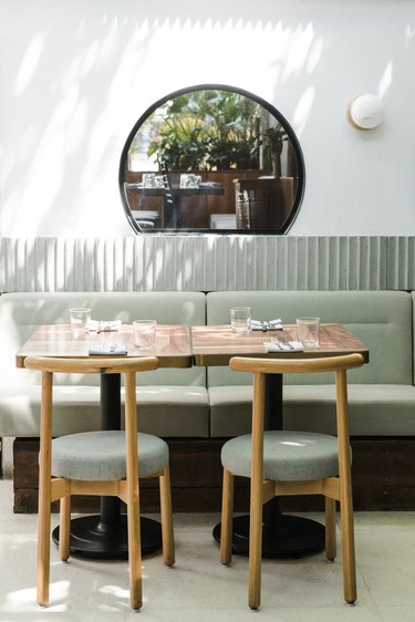 Restaurant area with curved wood chairs, wood tables, and green booths. Wall with a corrugated border, round window, and globe sconce.