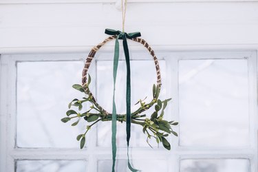 a finished diy mistletoe wreath with a green ribbon hangs from a ceiiing