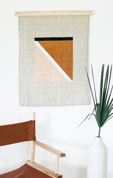 Painted quilt hanging with foldable brown chair and plant in vase