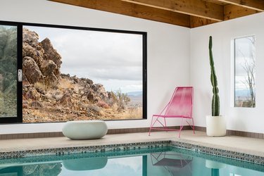 A pink metal chair with a cactus and large windows and a swimming pool