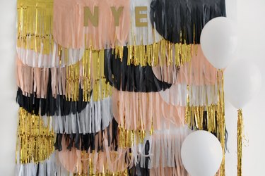 DIY NYE backdrop with scalloped paper fringes in black, white, pink, and gold, with white balloons in a white room