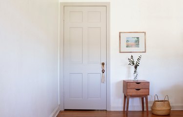 White walled room with wood flooring and wood side table, basket, vase, and art