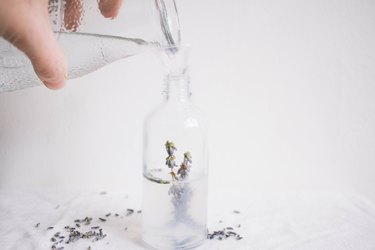 Water being poured from a clear pitcher into a spray bottle with herbs