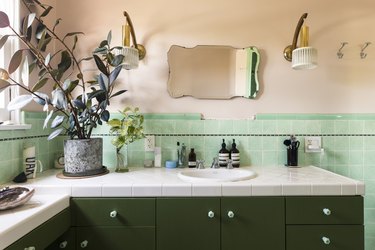 Bathroom vanity with a tiled counter, green cabinets, plants. The wall is green tiled and painted a peach color. Art deco sconces and a mirror.