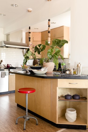 Red stool at a light wood kitchen island with black counter. Black and white dishes, a plant, and beaded pendant lights accent.