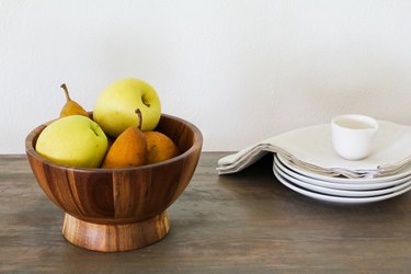 a wooden bowl full of fruit next to white saucers and napkins