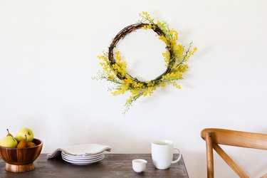 a wreath with tiny yellow flowers on a wall above a rustic wooden table set with white mugs and saucers