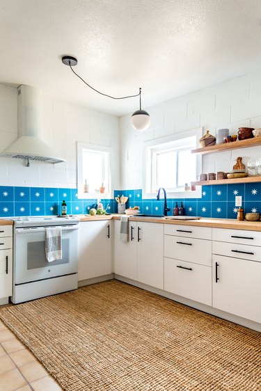 a kitchen with white cabinets and walls, a striking blue tile backsplash, and a jute rug