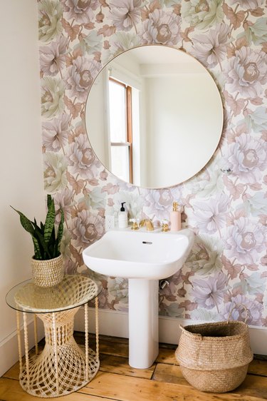 a bathroom with a pedestal sink, round mirror, and lavender-toned floral wallpaper