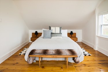 a white bedroom with a rustic wide-plank floor, a wooden bench at the foot of the bed, and matching built-in nightstands