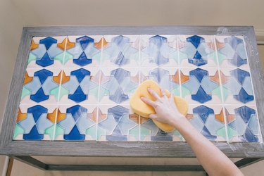 Person using a damp grout sponge to wipe away excess grout from a blue and orange tabletop, of a wood coffee table