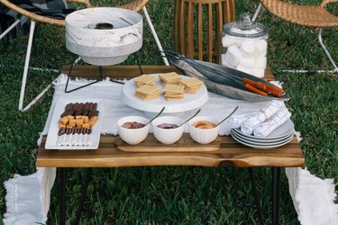 Outdoor s'mores picnic spread with a small fire pit, roaster sticks, and food ingredients
