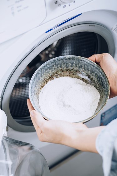 Hand holding bowl with white powder over white washing machine
