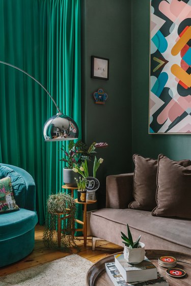 Corner of a living room featuring a chocolate brown couch, teal velvet chair, and emerald green walls and curtains.