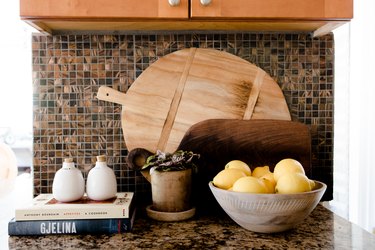 Granite kitchen counter with a bowl of lemons, cookbooks, wood cutting boards, and a brown tile backsplash.