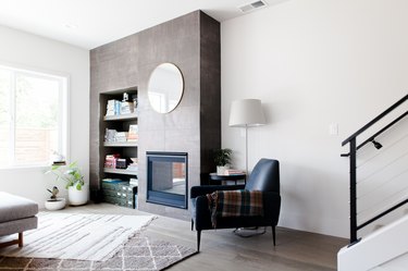 Living room with a gray accent wall, fireplace, round mirror, black armchair with a plaid blanket, layered rugs on a gray wood floor.