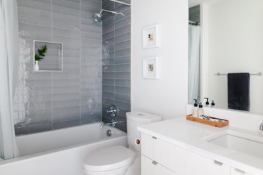 Bathroom with a white vanity, gray tile shower, hexagon floor tiles, and beige bath mats. Niche with a white vase and plant.