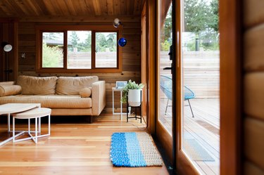 Living room with wood floors, wood walls, beige sofa, wood tables, planter stand, blue ornaments, casement door, and blue-beige doormat.