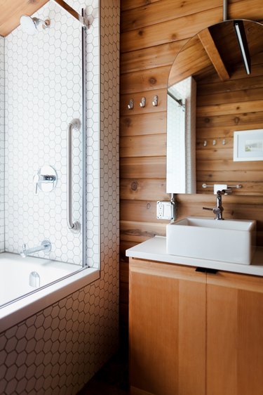 A bathroom with a wood vanity and wood walls. A square sink basin and an arched mirror. A shower with white hexagon tiles.