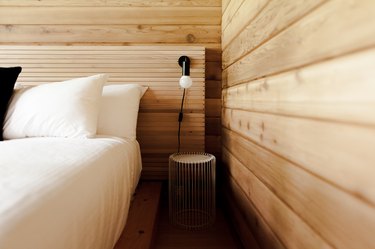 A wood headboard, white bedding, and wood walls. A black wall sconce and a round, white wire night table.