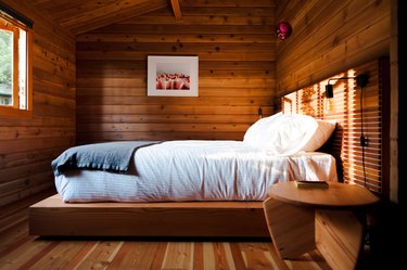 A bedroom with wood walls, wood ceiling, wood bed frame, and wood table. Black lamp wall sconces and white-gray bedding.