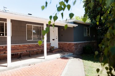 A brick and concrete walkaway to a gray house with brick siding and wood benches.