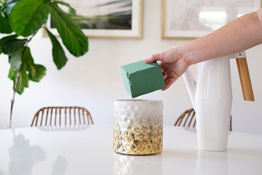 Hand putting a block of green floral oasis in a textured gold-white vase