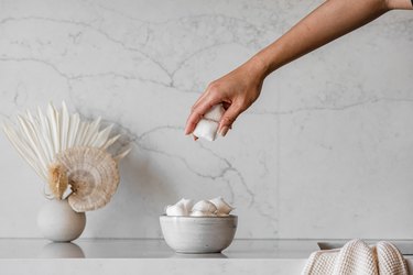 A person taking a detergent pod from a bowl on a granite or marble backsplash countertop with a vase of dried florals