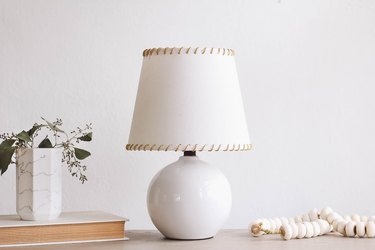 SKOTTORP lampshade with leather whipstitching, on a table with a book, and a vase of flowers