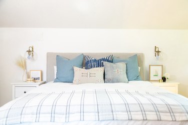 Linen upholstered headboard of a bed with blue and gray pillows, and double white nightstands and globe sconces