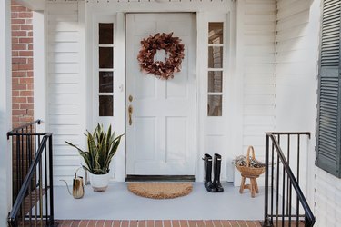 A white traditional door with side lights and a fall wreath; a plant, watering can, boots, and a basket are on the front porch, which has black railings